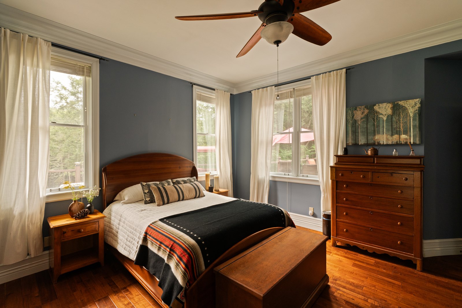 A bedroom with a wooden bed frame, blue walls and windows on two sides.
